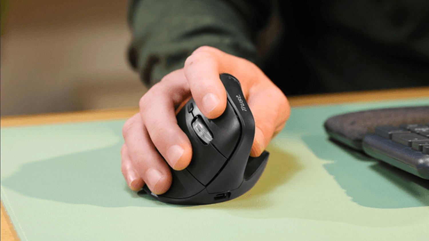 Pravá ruka používá černou ergonomickou myš Trust Vyran na zelené podložce vedle klávesnice.