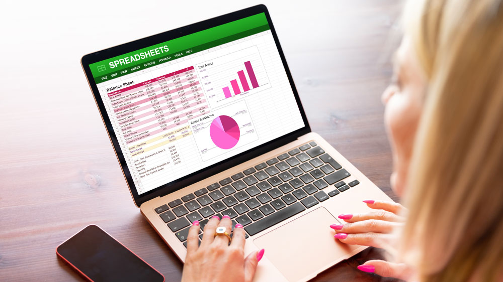 Mladá žena zpracovává tabulky v programu Excel na notebooku. Vedle notebooku je položený mobilní telefon_02