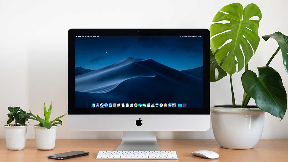 Monitor Apple s bezdrátovou klávesnicí, myší a mobilním telefonem stojícím na stole mezi květináči se zelenými rostlinami_02