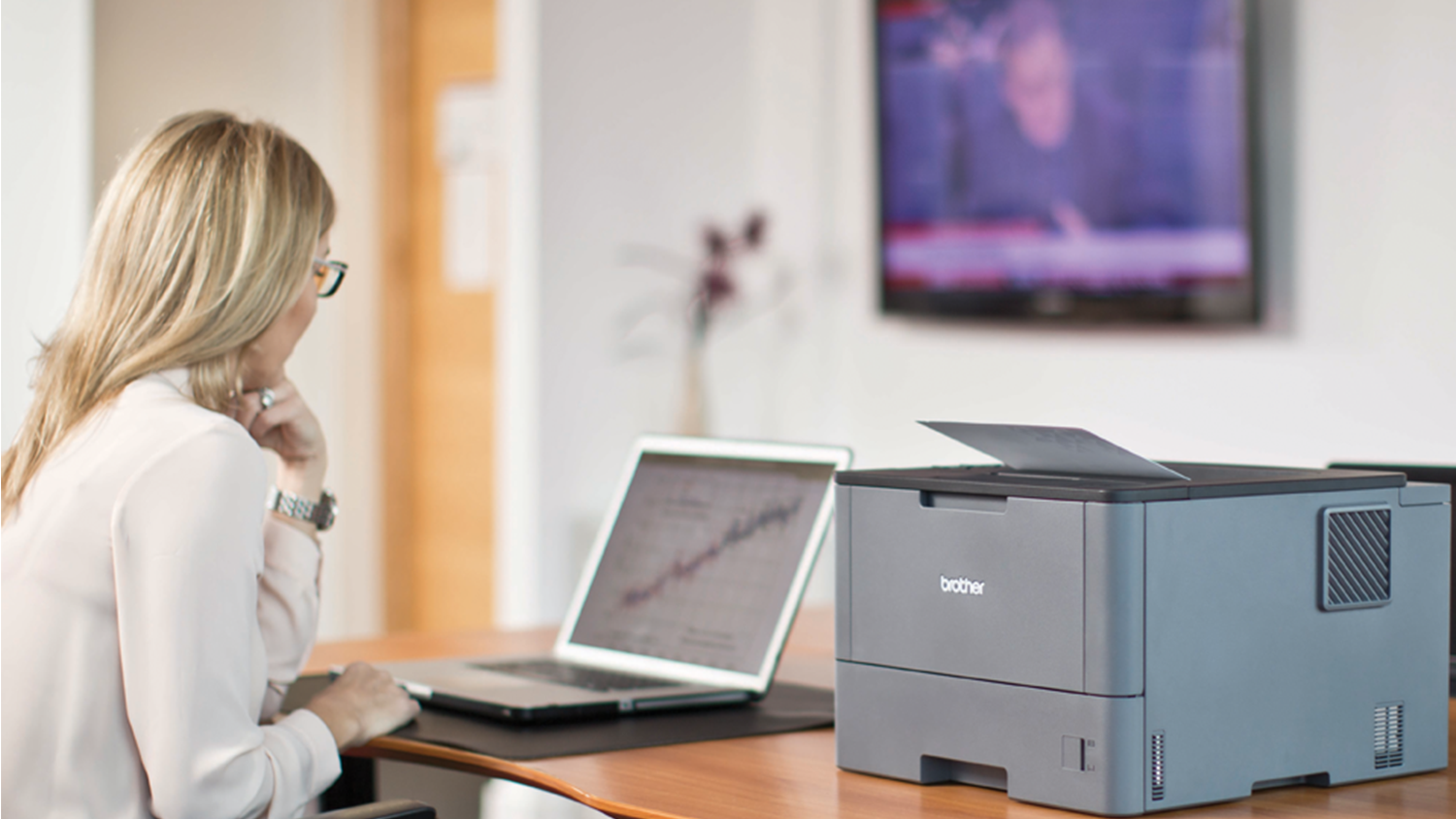 Žena sed&iacute; pri stole a d&iacute;va sa do notebooku, ktor&yacute; lež&iacute; na stole vedľa laserovej tlačiarne Brother HL-L5000D_02