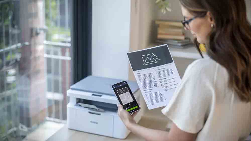 Žena u laserov&eacute; tisk&aacute;rny Brother DCP-L1630W s pap&iacute;rem a mobilem v ruce_02