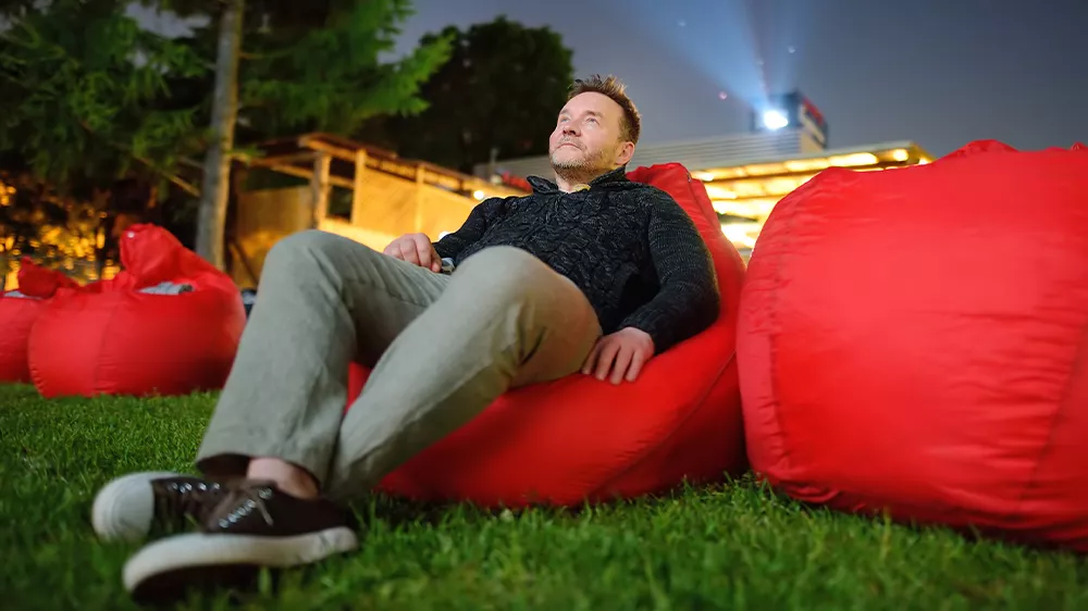 Muž sed&iacute;c&iacute; na červen&eacute;m pol&scaron;t&aacute;ři a sleduj&iacute;c&iacute; film, kter&yacute; se prom&iacute;t&aacute; z projektoru Dangbei Mars Pro 2.