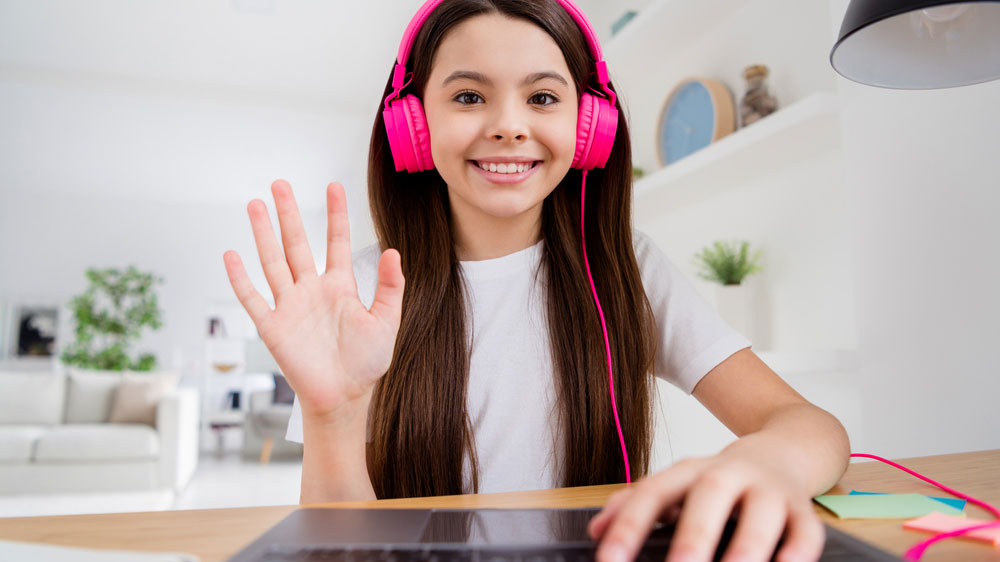 Děvče s růžovými sluchátky mává do kamery notebooku