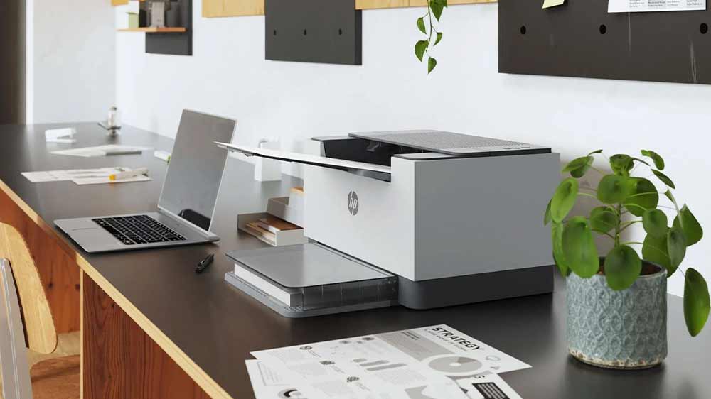 Printer HP LaserJet M209d na stole v kanceláři vedle květiny a notebooku_03