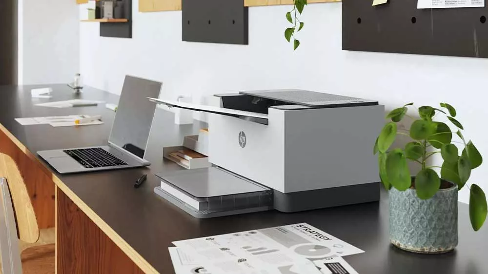 Printer HP LaserJet M209d na stole v kanceláři vedle květiny a notebooku_03