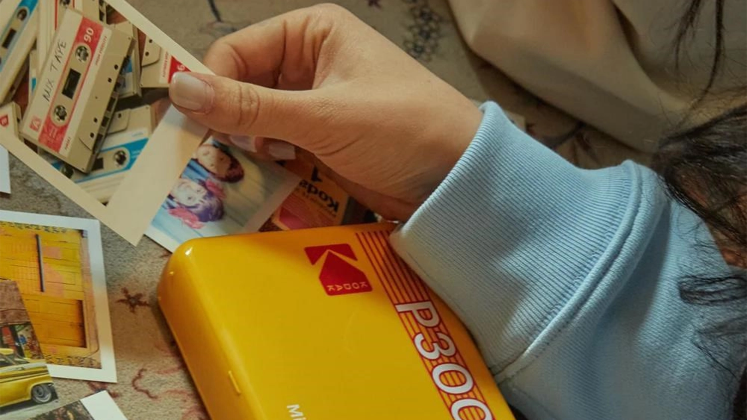 Tisk&aacute;rna Kodak s fotografiemi na posteli a ruka ženy drž&iacute;c&iacute; sn&iacute;mek