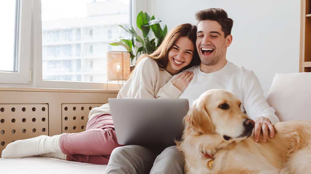Smějící se mladý pár sedí v obýváku na sedačce a sleduje vtipná videa na notebooku Lenovo ThinkBook 16 G7 ARP. Vedle nich leží zlatý retrívr_03