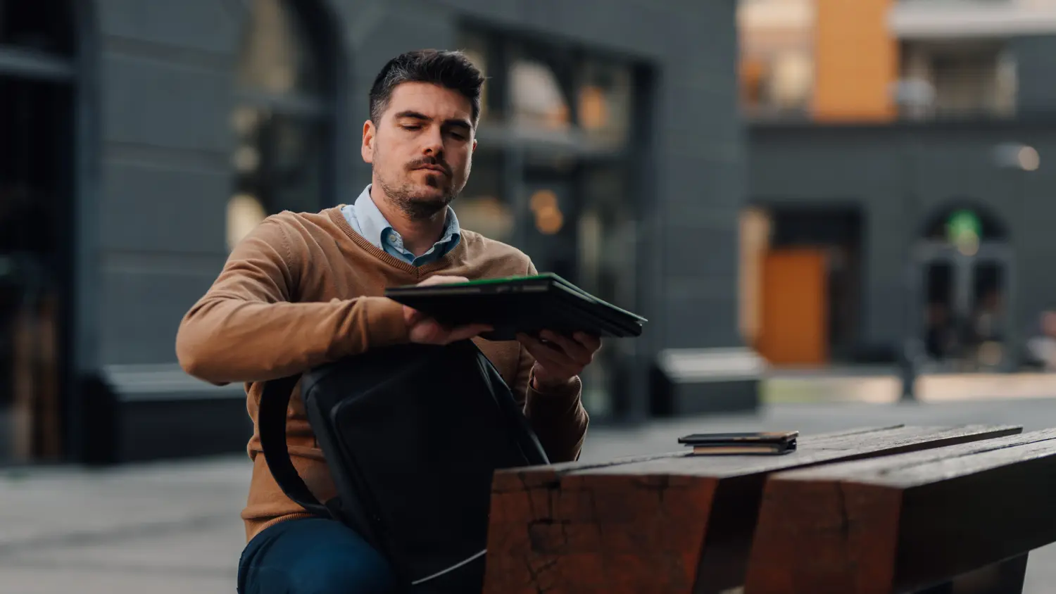 Muž sedí vonku pri stole a vyberá z batoha notebook