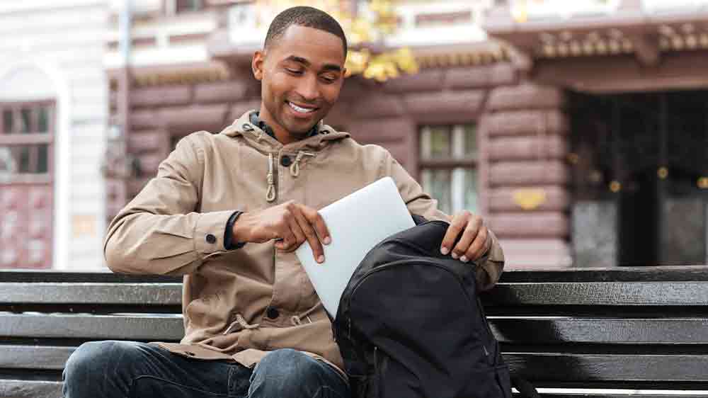 Mladý černý muž sedící na lavičce a ukládající notebook do batohu_02