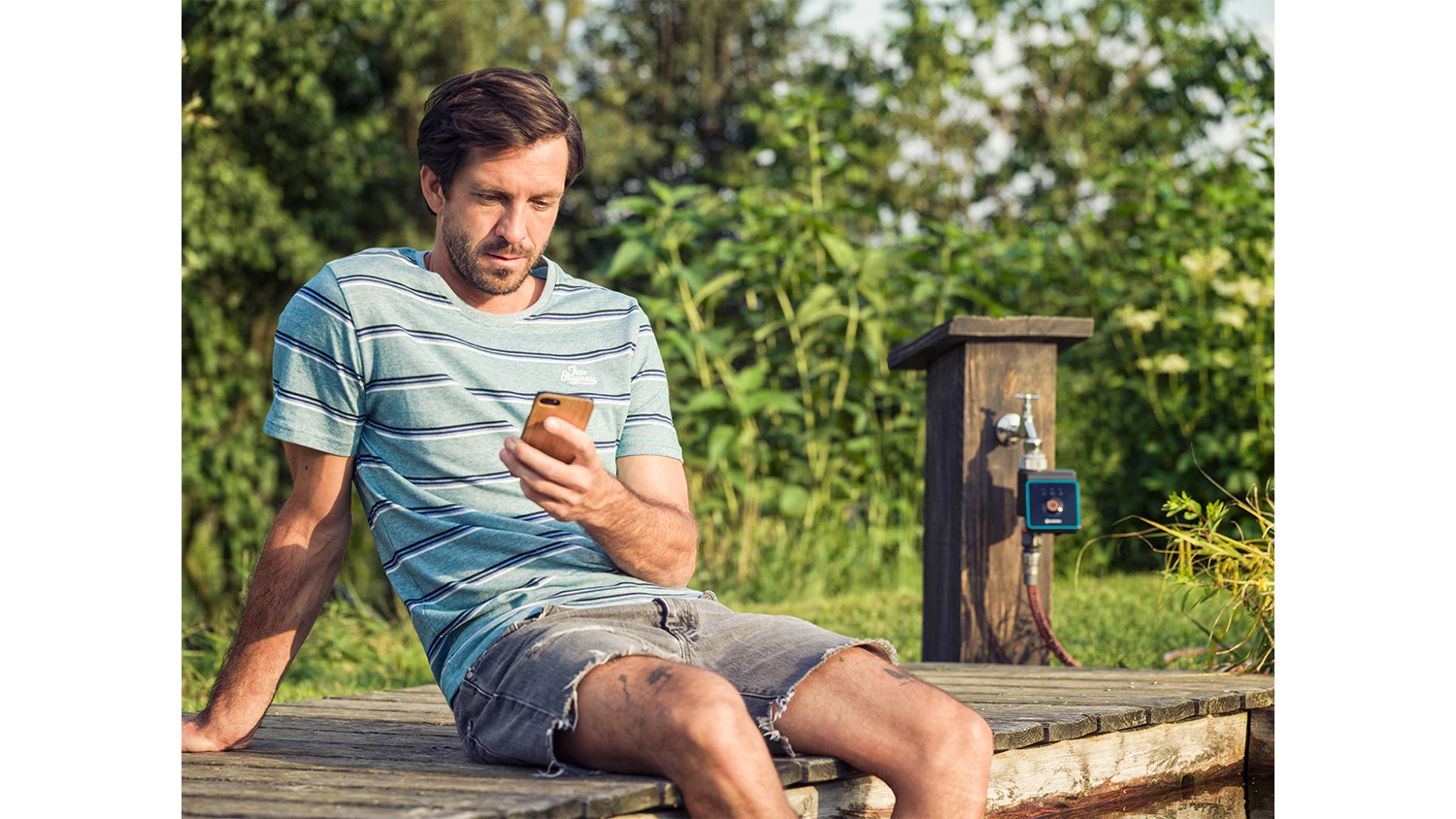Muž s mobilem v ruce, v pozad&iacute; zavlažovac&iacute; poč&iacute;tač GARDENA Bluetooth