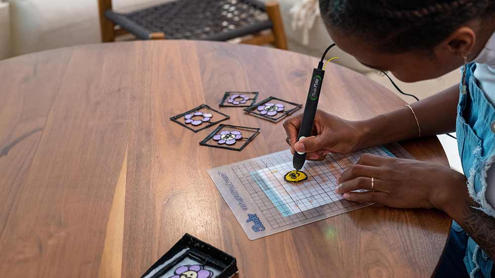 Žena sedící u stolu a tvořící obrázek 3D perem 3Doodler FLOW+ na podložce se šablonou_06