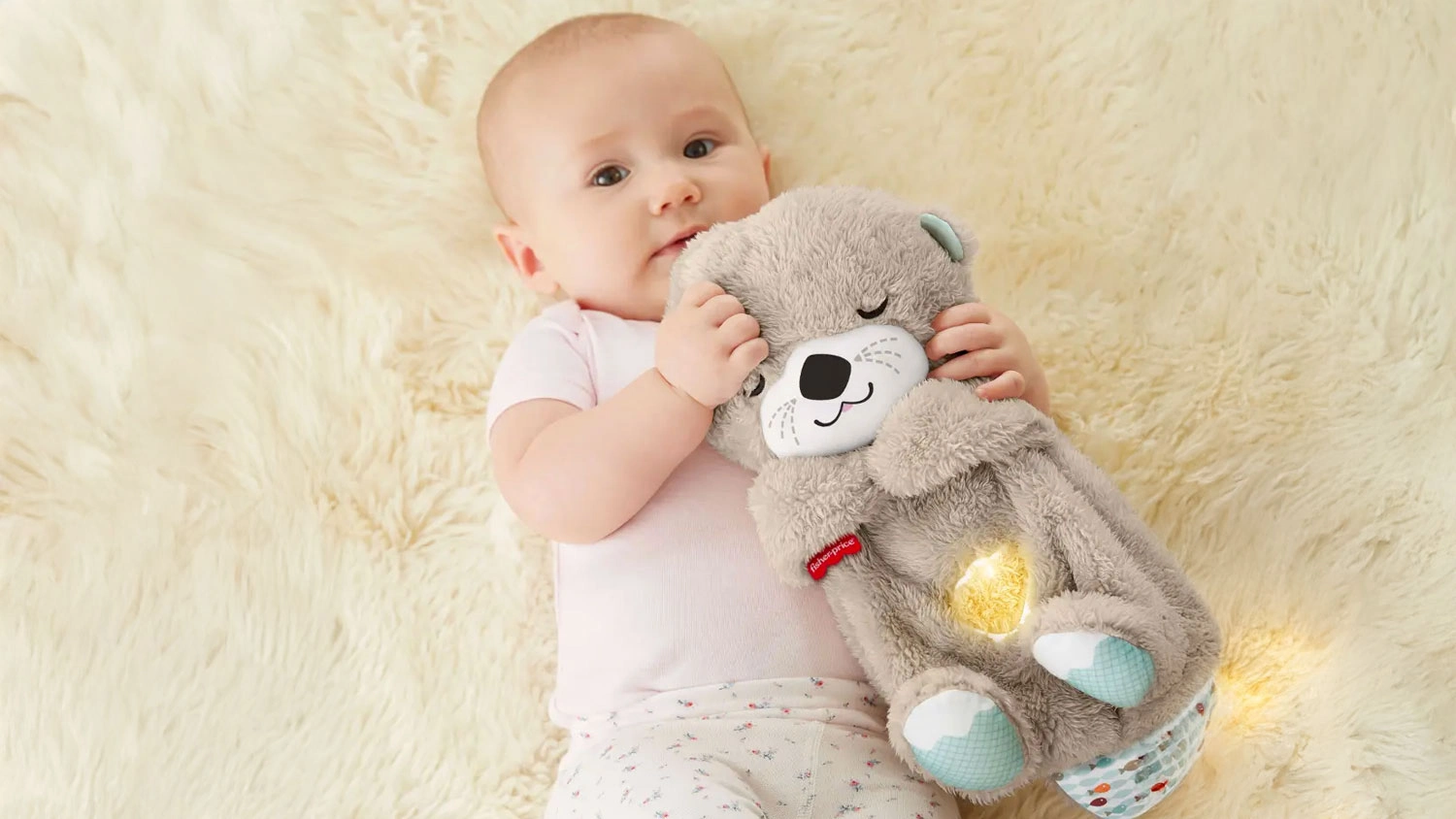 Miminko lež&iacute; na hebk&eacute; kože&scaron;ině a obj&iacute;m&aacute; ply&scaron;ovou zklidňuj&iacute;c&iacute; vydru Fisher-Price s jemně sv&iacute;t&iacute;c&iacute;m bř&iacute;&scaron;kem.