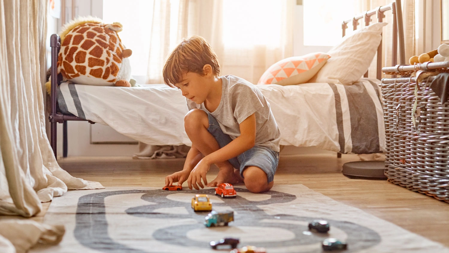 Malý chlapec klečí v dětském pokoji na koberci s motivem dráhy a hraje si s autíčky Hot Wheels.
