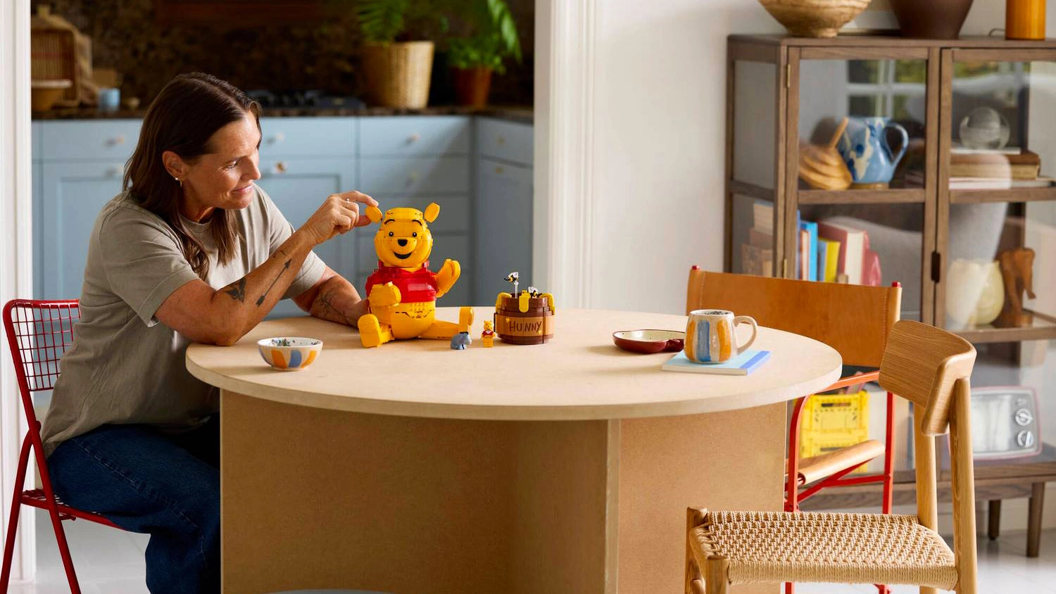 Usmívající se žena sedí u stolu a polohuje figurku ze stavebnice LEGO® | Disney 43300 Medvídek Pú