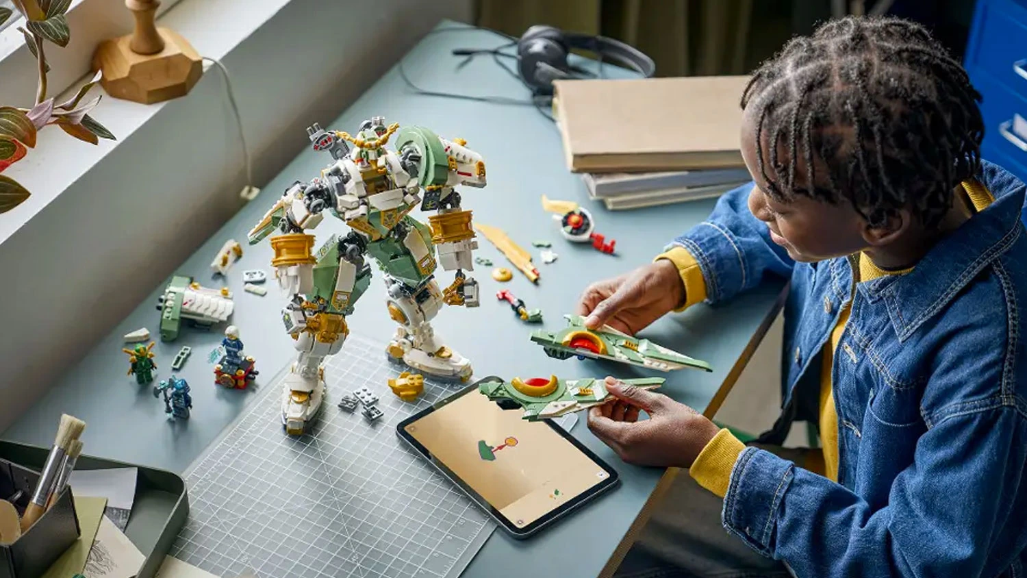 Usmívající se chlapec sestavuje stavebnici LEGO® NINJAGO® 71860 Lloydův titanový robot: 15 let NINJAGO® a v tabletu má otevřený návod v aplikaci LEGO® Builder