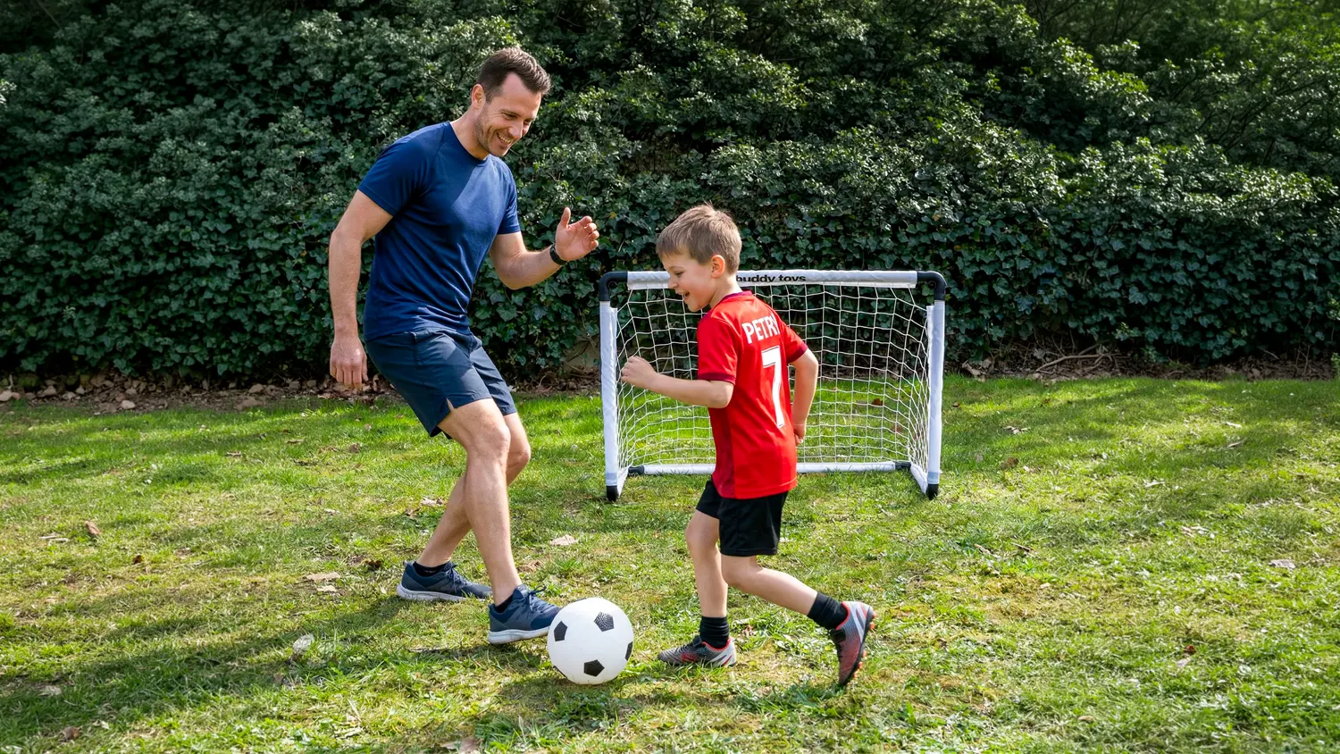 Muž a chlapec hrají společně fotbal na tzrávě před brankou Buddy toys.