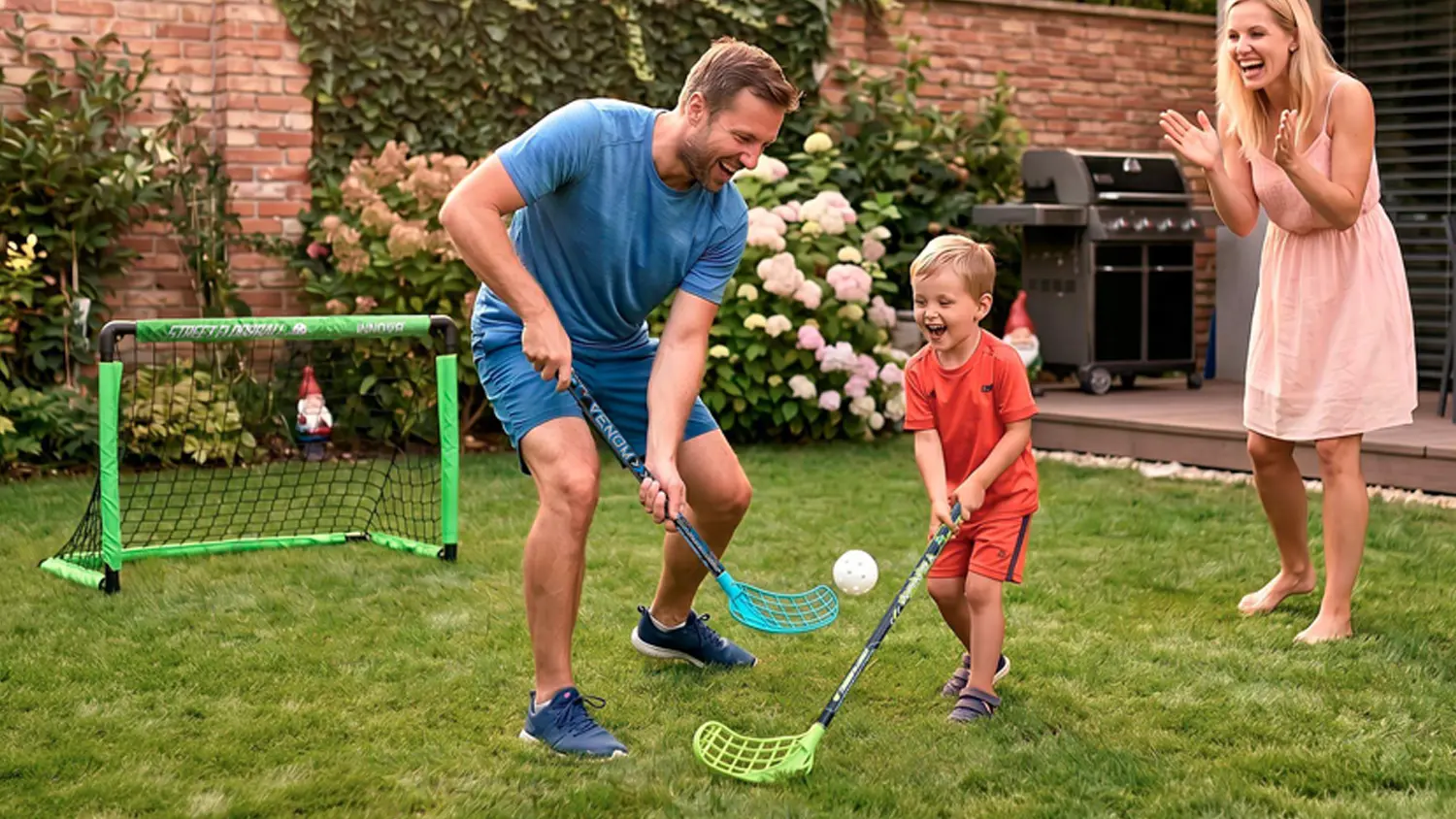 Muž s chlapcem hrají florbal na trávě před domem před brankou Buddy Toys, zatímco žena jim fandí.