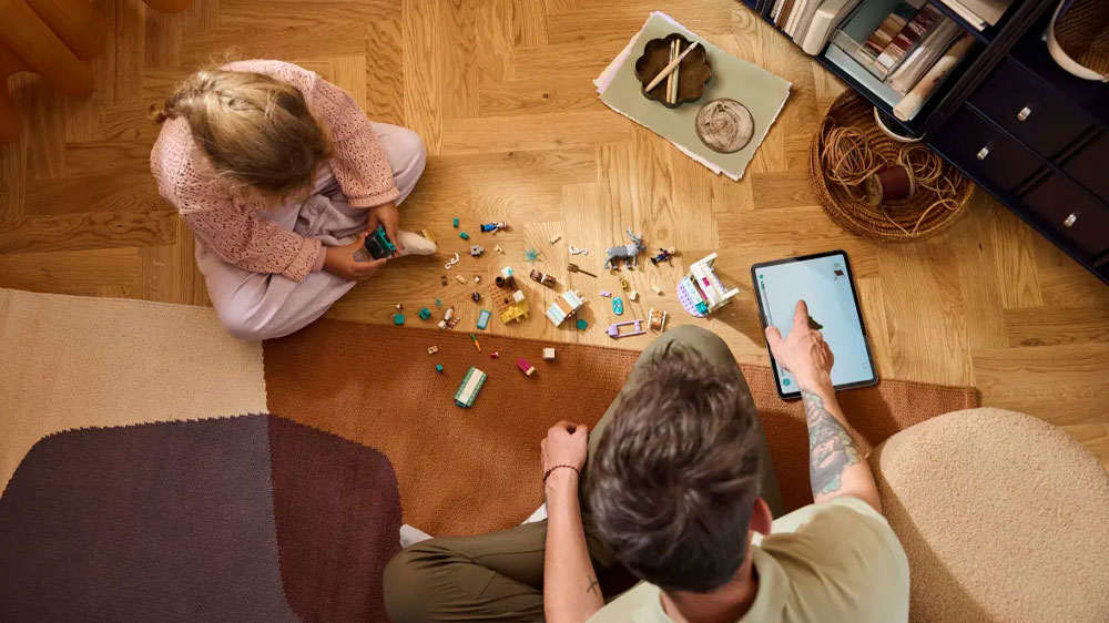 Dcera s tatínkem staví stavebnici LEGO® Dysney Princess 43256 Anna a dobrodružství na saních za pomoci aplikace Lego Builder na zemi v pokoji_02