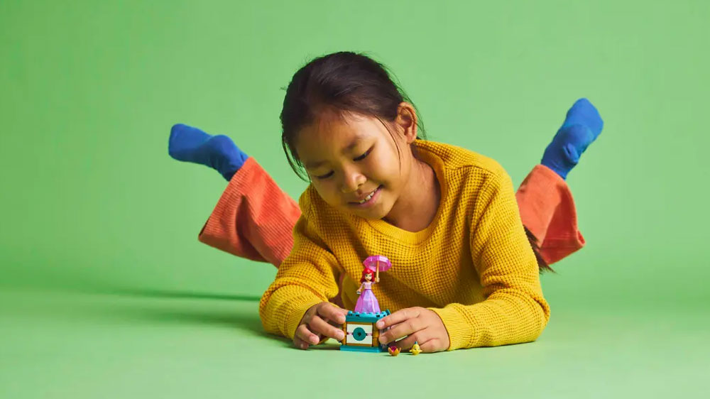 Holčička ležící na zemi na břiše a hrající si se stavebnicí LEGO® Dysney Princess 43259 Točící se Ariel na zeleném pozadí a podkladu_03