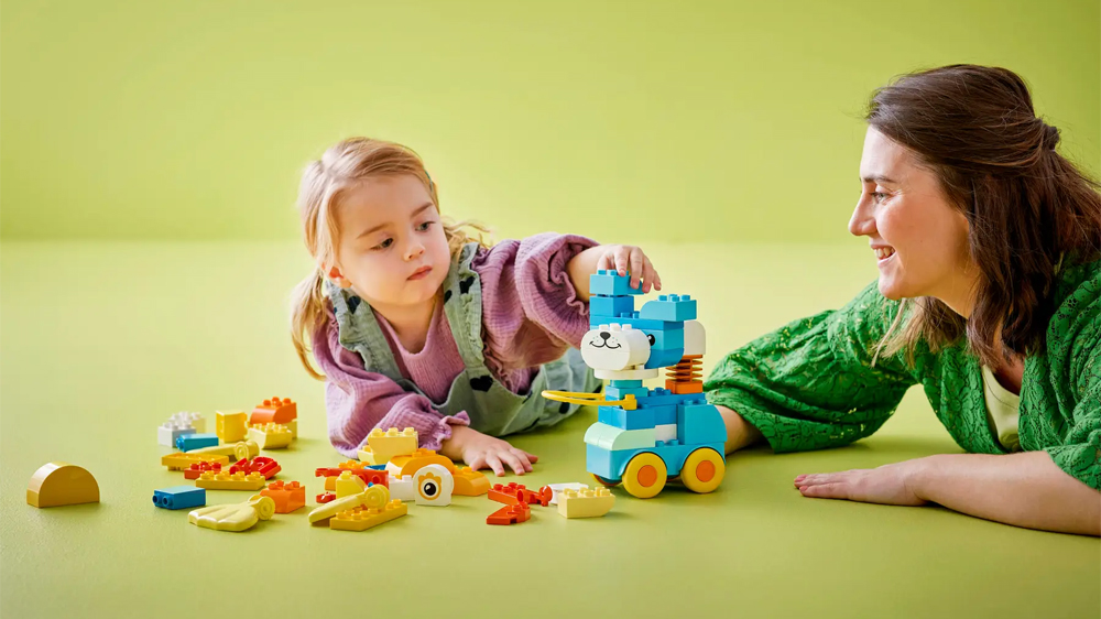 Žena se usm&iacute;v&aacute; na holčičku, kter&aacute; skl&aacute;d&aacute; pejska ze stavebnice LEGO&reg; DUPLO&reg; 10448 3 v 1: Zv&iacute;ř&aacute;tka na kolečk&aacute;ch_02