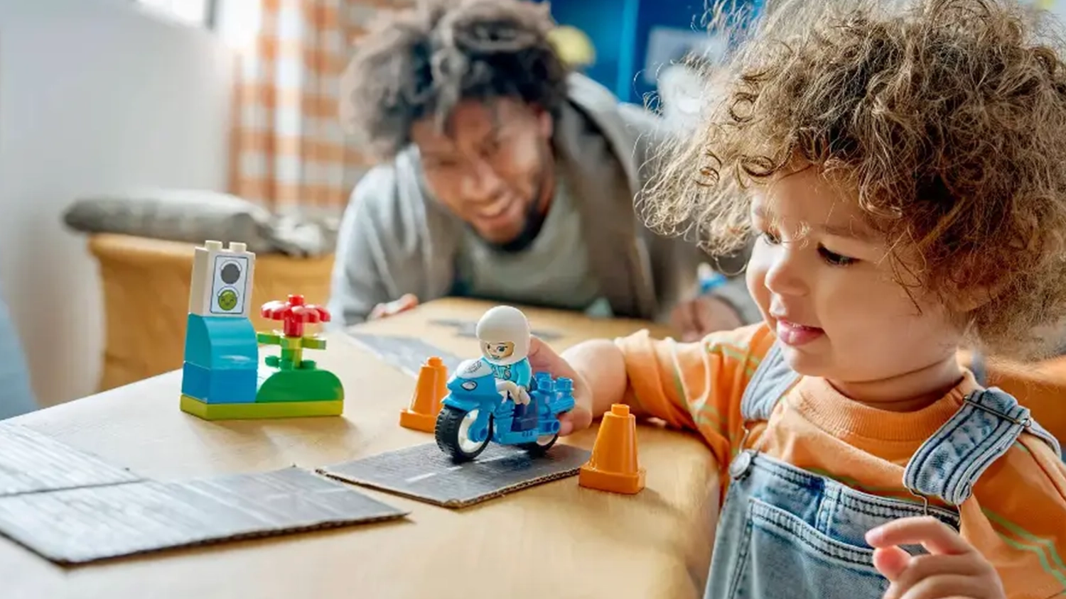 Otec se usm&iacute;v&aacute; na mal&eacute;ho chlapce, kter&yacute; jezd&iacute; s figurkou policistky ze stavebnice LEGO&reg; DUPLO&reg; (10471) mezi kužely.