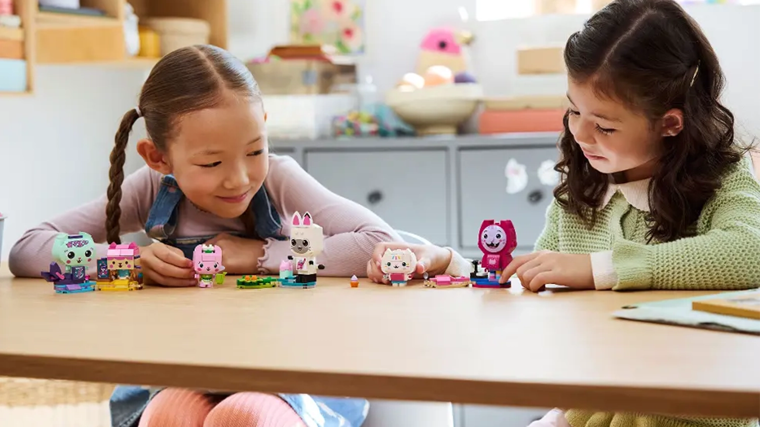Dve dievčatá sedia pri stole a hrajú sa s figúrkami zo stavebnice LEGO® Gabby’s Dollhouse 11215 Gabby a mačací kamaráti z kociek.