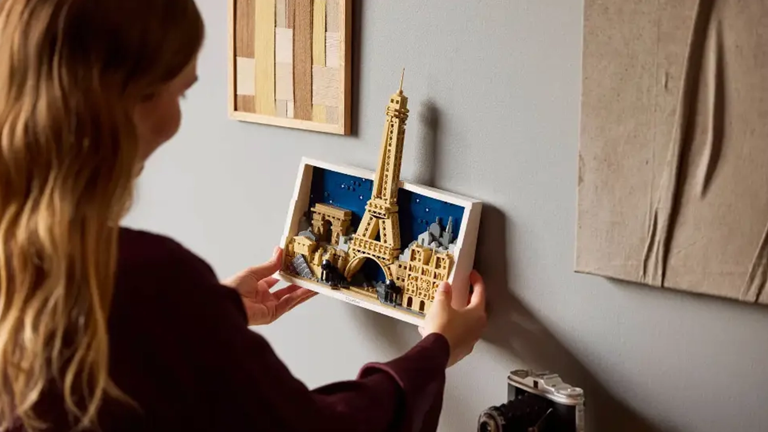 Žena přidělává na zeď dekoraci v rámečku ze stavebnice LEGO® Architecture 21064 Paříž – město lásky.