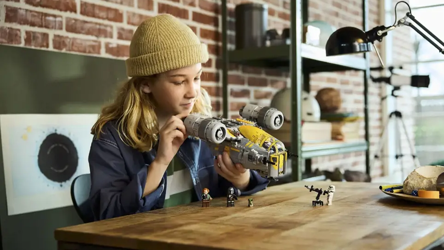 Dívka sedící u stolu si hraje s replikou vesmírné lodi Razor Crest™ ze stavebnice LEGO® Star Wars™ (75447).