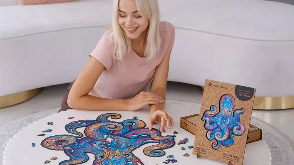 Usmívající se dívka skládá dřevěné puzzle Unidragon Magneti Octopus na bílém konferenčním stolku v obývacím pokoji, za ní je bílá pohovka s plyšovou chobotnicí_03