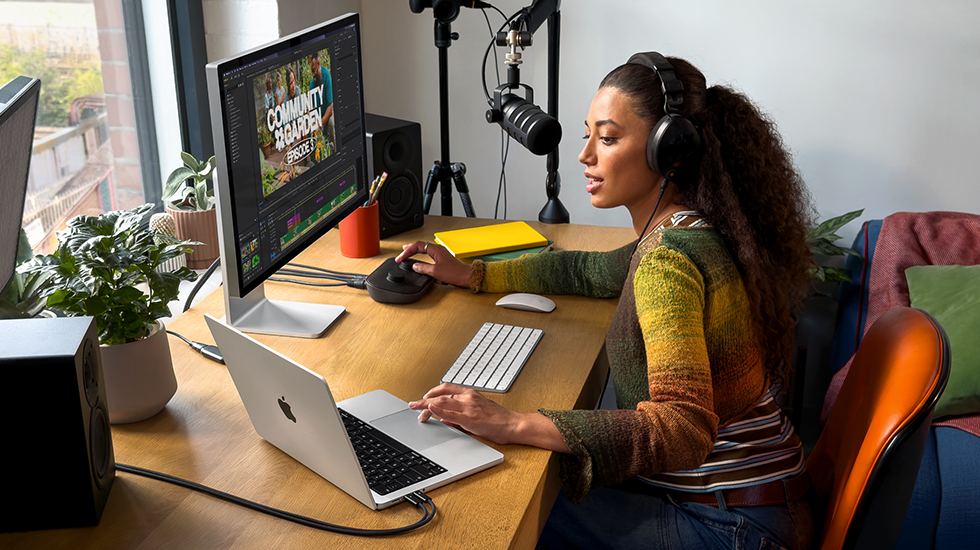 Žena sedí doma u&nbsp;pracovního stolu a&nbsp;používá MacBook&nbsp;Pro připojený k&nbsp;externímu monitoru, má nasazená sluchátka a&nbsp;mluví do mikrofonu