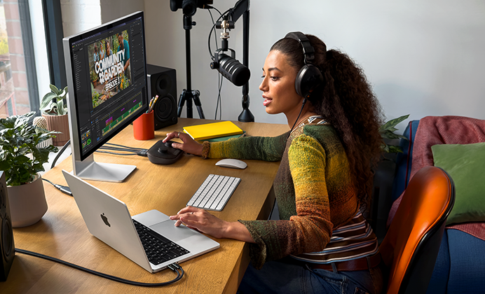 Žena sedí doma u&nbsp;pracovního stolu a&nbsp;používá MacBook&nbsp;Pro připojený k&nbsp;externímu monitoru, má nasazená sluchátka a&nbsp;mluví do mikrofonu