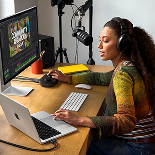 Žena sedí doma u&nbsp;pracovního stolu a&nbsp;používá MacBook&nbsp;Pro připojený k&nbsp;externímu monitoru, má nasazená sluchátka a&nbsp;mluví do mikrofonu