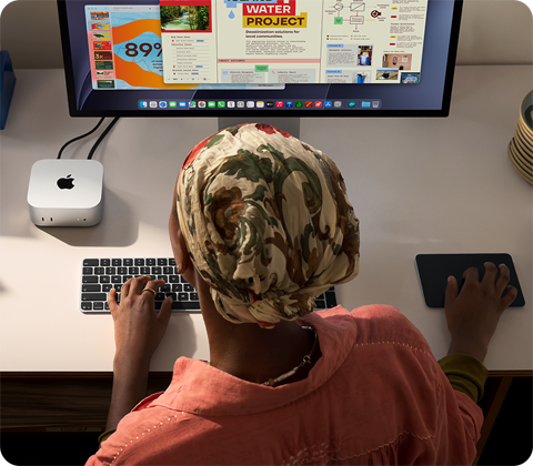 Osoba píše u stolu na Macu mini připojeném ke Studio Displayi, Magic Keyboard, Magic Trackpad, různé předměty