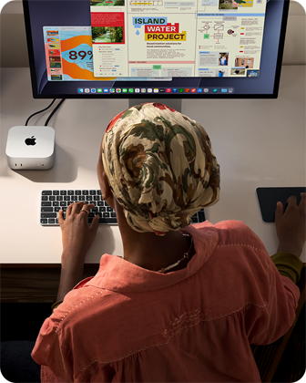 Osoba píše u stolu na Macu mini připojeném ke Studio Displayi, Magic Keyboard, Magic Trackpad, různé předměty