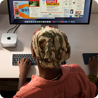 Osoba píše u stolu na Macu mini připojeném ke Studio Displayi, Magic Keyboard, Magic Trackpad, různé předměty