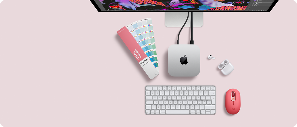 Pohled shora na Mac mini, stříbrný hliník, zaoblené rohy, uprostřed lesklé černé logo Apple, připojení kabelem, nahoře je částečně vidět Studio Display, otevřené AirPody 4, vzorky barev Pantone, číselná klávesnice a myš