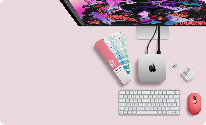 Pohled shora na Mac mini, stříbrný hliník, zaoblené rohy, uprostřed lesklé černé logo Apple, připojení kabelem, nahoře je částečně vidět Studio Display, otevřené AirPody 4, vzorky barev Pantone, číselná klávesnice a myš