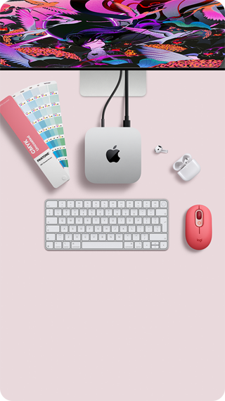 Pohled shora na Mac mini, stříbrný hliník, zaoblené rohy, uprostřed lesklé černé logo Apple, připojení kabelem, nahoře je částečně vidět Studio Display, otevřené AirPody 4, vzorky barev Pantone, číselná klávesnice a myš